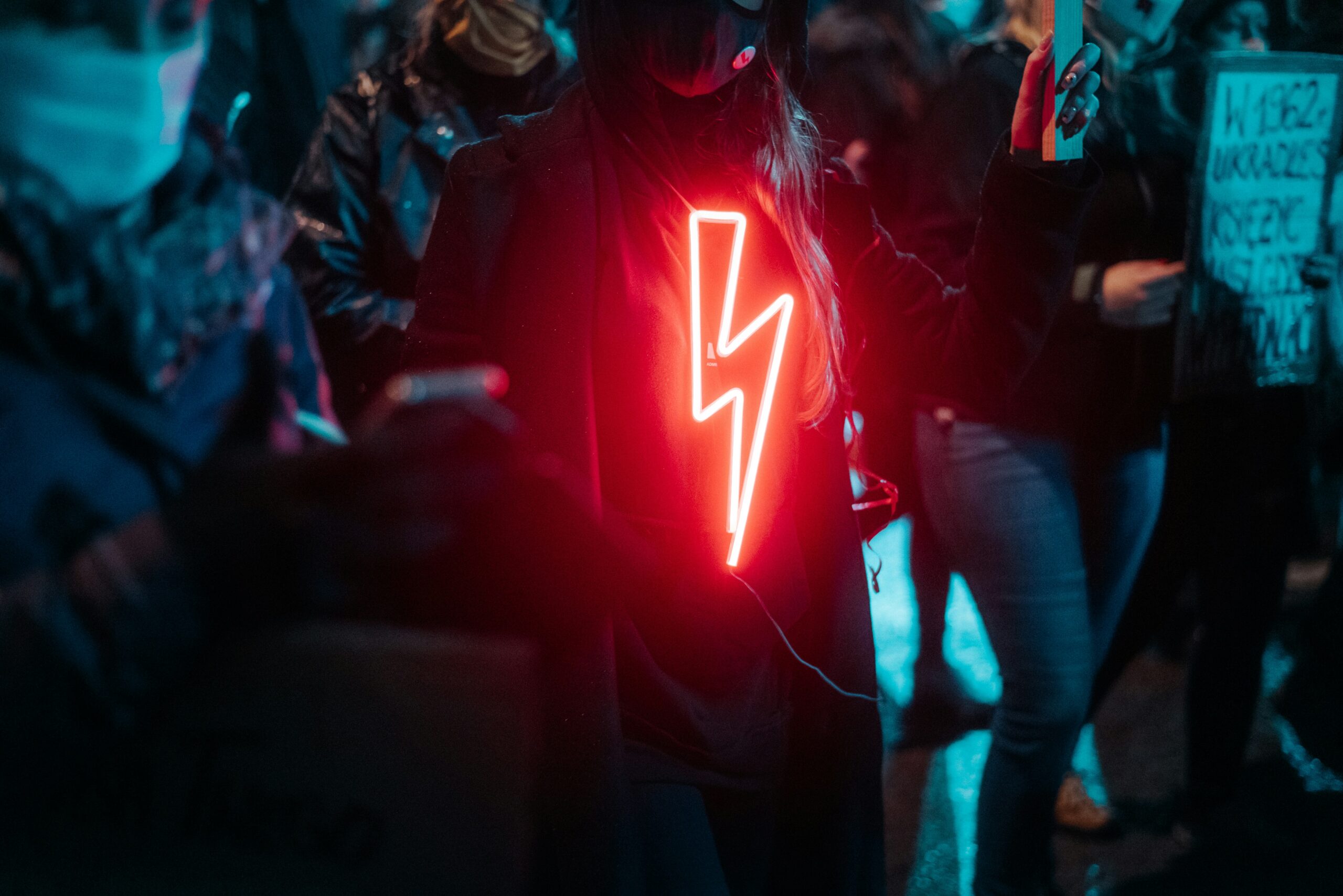 Lightning bolt neon sign on person's black jacket at night, illuminated in red, in a crowd of people.