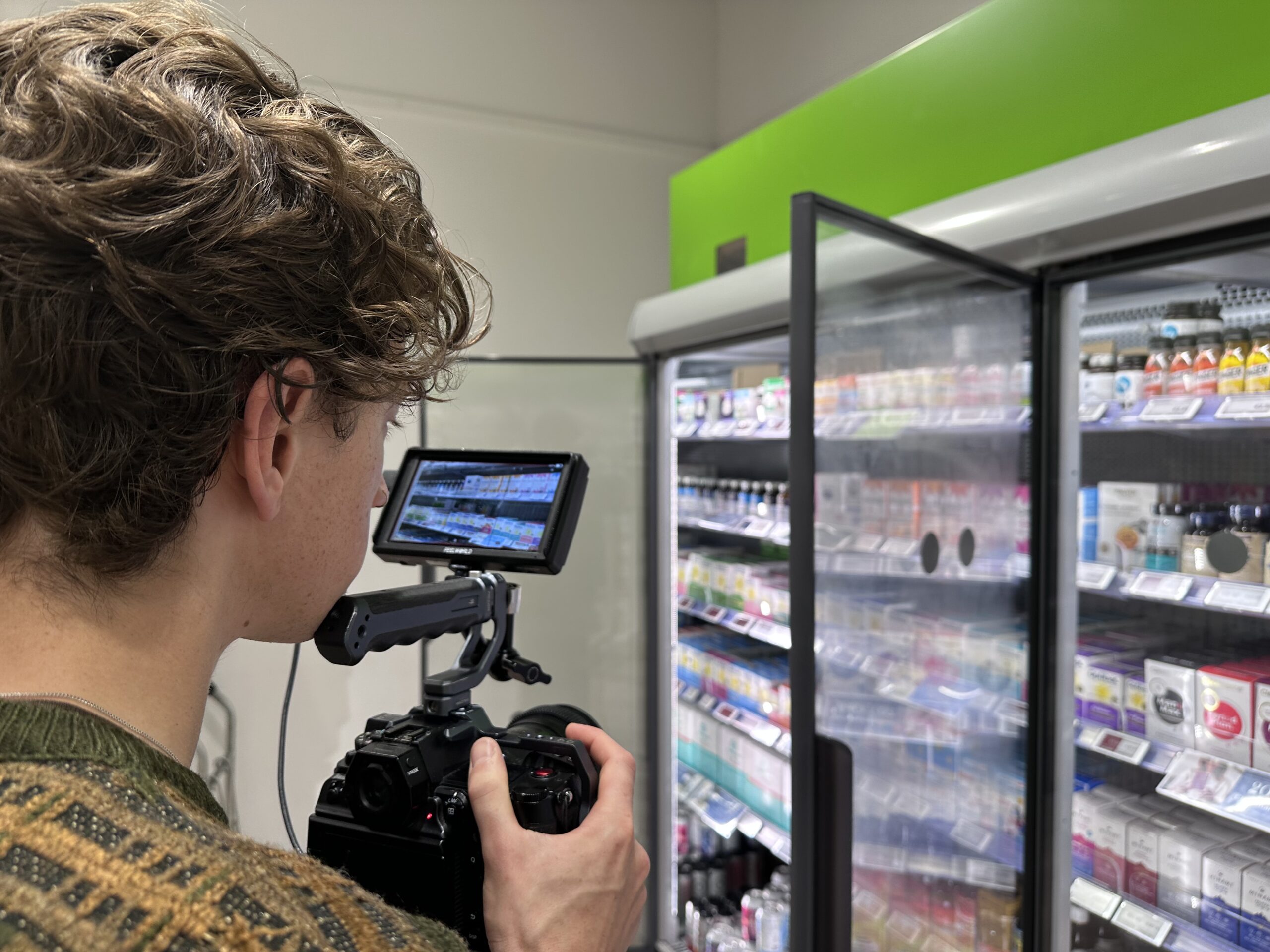 Focused young person photographing product display at supermarket using professional camera and stabiliser, capturing detailed images of dairy products in a refrigerated section for marketing or review purposes.