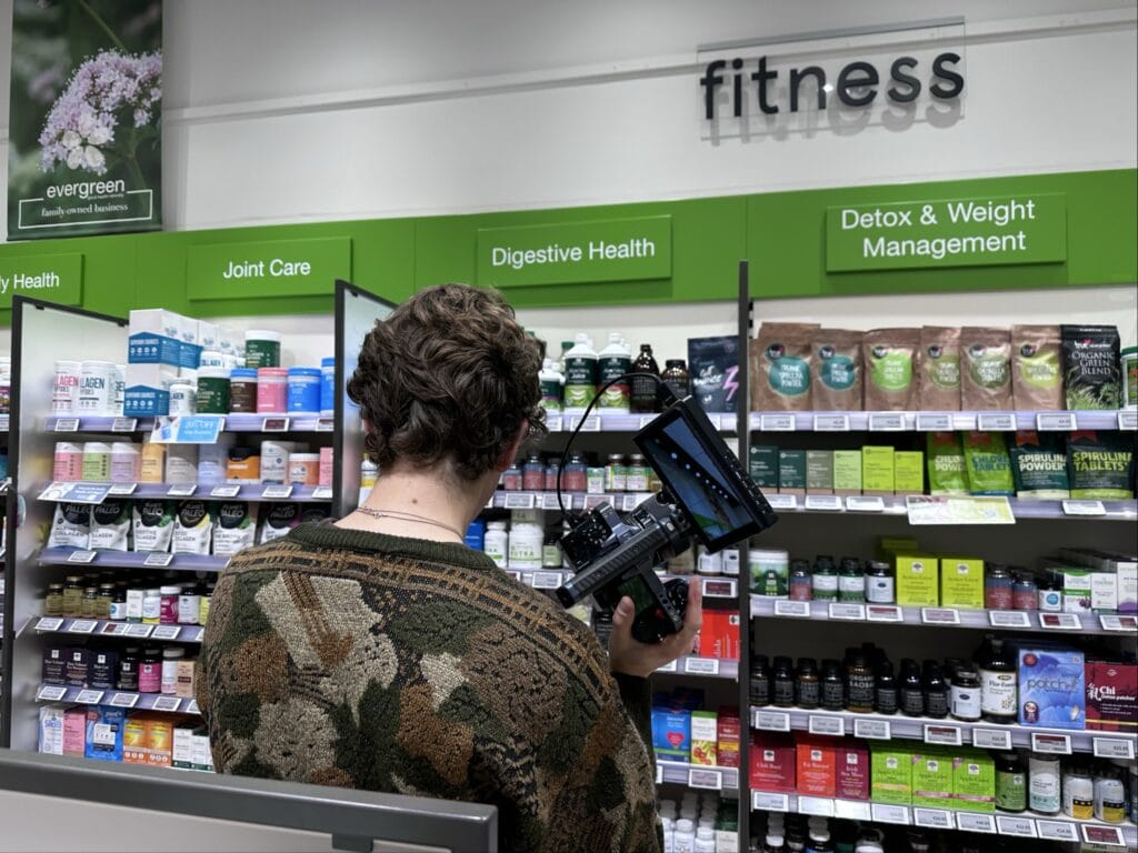 Image 12 | mindboost. Vibrant health supplement aisle with a person filming or photographing nutritional products at a health store, showcasing dietary supplements, vitamins, and herbal remedies, promoting overall wellness and health consciousness.